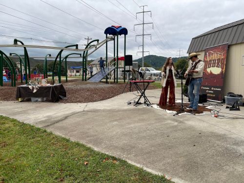 A musician and singer perform at a park event near playground equipment and a decorated table.