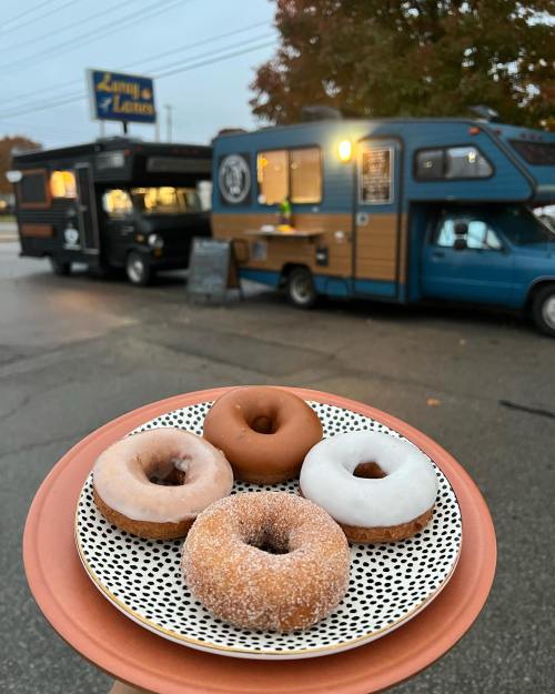 A plate of four donuts in front of a food truck, with a cozy autumn setting and colorful trees in the background.