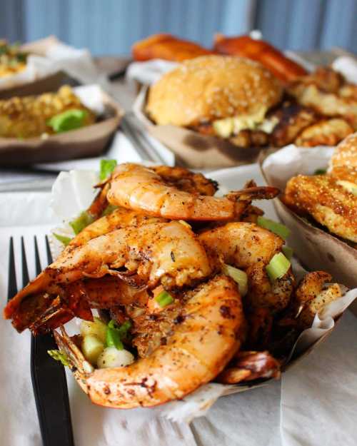 A close-up of seasoned shrimp garnished with green onions, with burgers and other dishes blurred in the background.