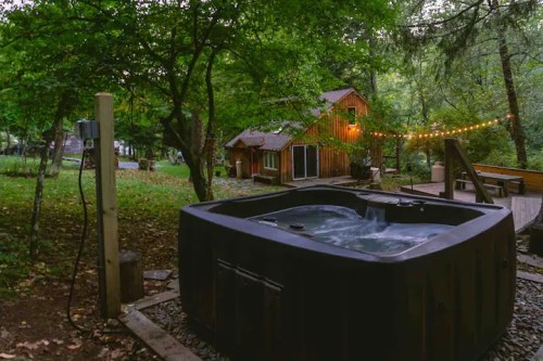 A hot tub surrounded by trees, with a cozy cabin and string lights in the background, creating a serene outdoor setting.