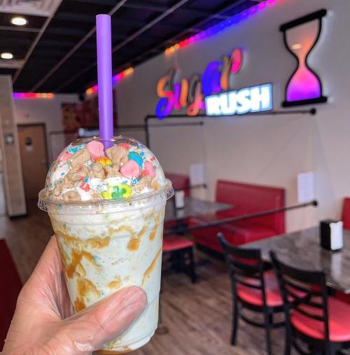 A hand holds a colorful milkshake topped with candy, with a vibrant "Sugar Rush" sign in the background.