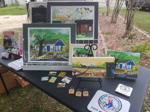 A display of colorful paintings and art pieces on a table, featuring houses, landscapes, and local scenes.