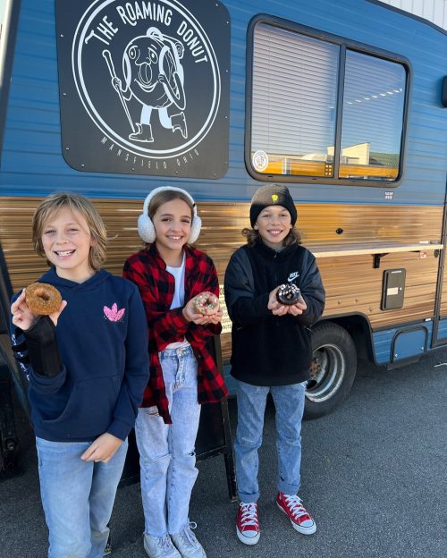 Three kids stand in front of a food truck, each holding a donut, smiling in casual winter attire.