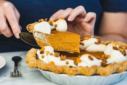 A person slices into a pumpkin pie topped with whipped cream and nuts, ready to serve a piece.