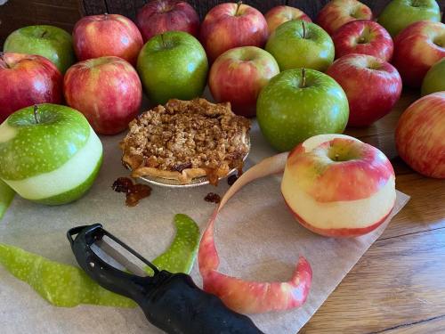 A variety of red and green apples surround a small apple pie, with an apple peeler and apple peels on a countertop.