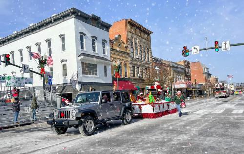 A festive parade scene with a decorated float, Santa, and a Jeep driving down a snowy street lined with buildings.