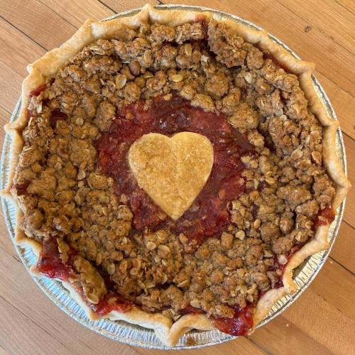 A pie with a crumbly topping and a heart-shaped cutout in the center, resting in a silver pie tin on a wooden surface.