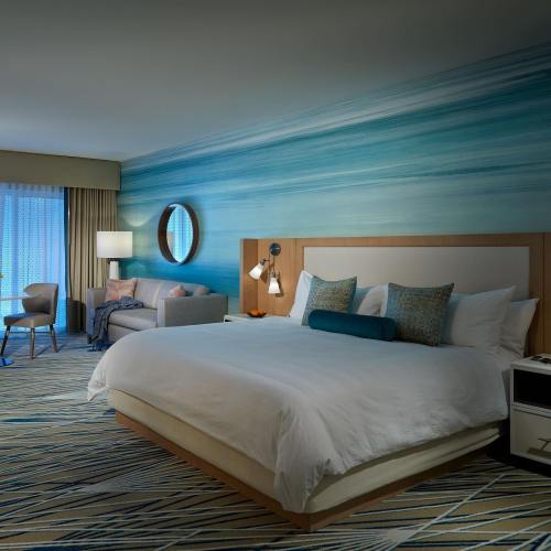 Modern hotel room featuring a king bed, blue accent wall, seating area, and large window with natural light.
