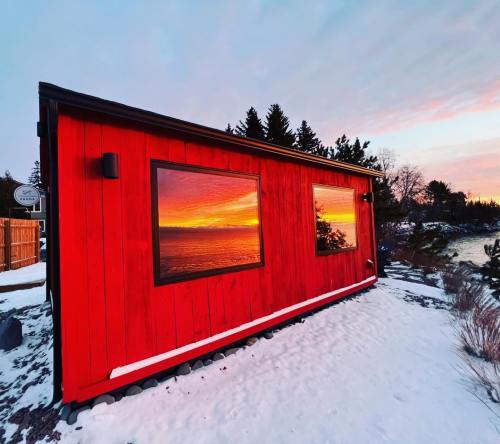 A vibrant red cabin reflects a colorful sunset over water, surrounded by snow and trees.