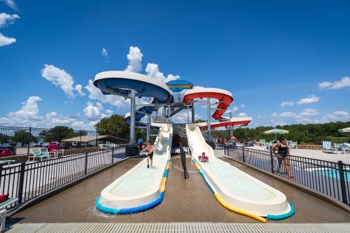Two water slides at a pool, with people enjoying the slides under a bright blue sky and fluffy clouds.