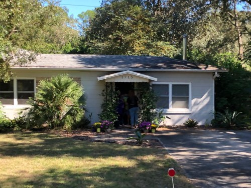 A small, light-colored house with a front porch, surrounded by greenery and flowers, on a sunny day.