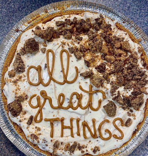 A pie topped with cream, crushed chocolate, and the words "all great things" written in caramel.