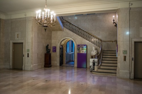 Elegant interior of a building featuring a staircase, chandelier, and sculptures, with marble floors and soft lighting.