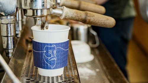 A coffee cup with a blue sleeve featuring a bearded face design, placed on an espresso machine.