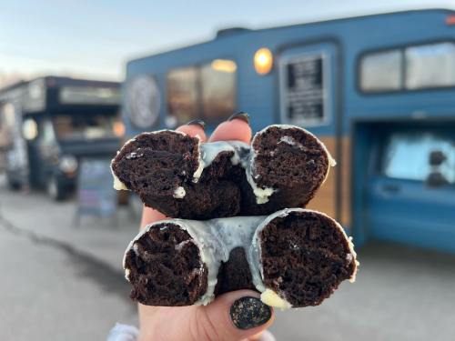 A hand holds four chocolate cookie rolls drizzled with white icing, with food trucks in the background.