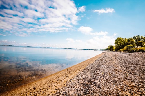 A serene lakeside view with a sandy shore, calm water, and a clear blue sky dotted with clouds.