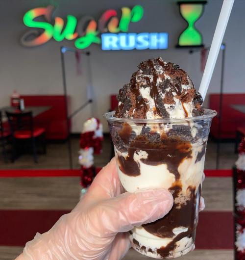 A hand in a glove holds a dessert cup with layers of ice cream, chocolate sauce, and crushed cookies, with a colorful sign in the background.