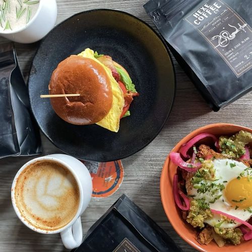 A plate with a burger, a bowl of salad, a cup of latte, and coffee bags on a wooden table.
