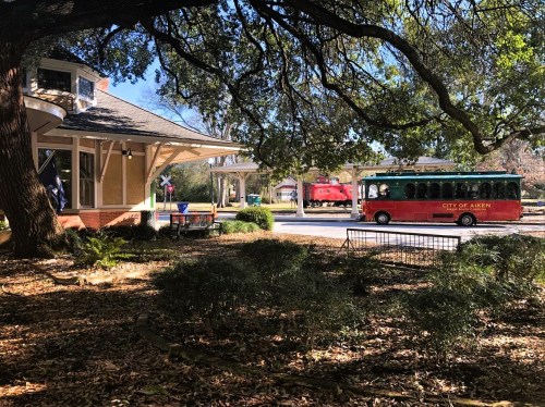 A red and green trolley bus passes by a charming building under large trees in a sunny park setting.