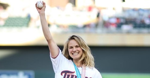A smiling woman in a Minnesota Twins jersey raises a baseball in the air at a baseball stadium.