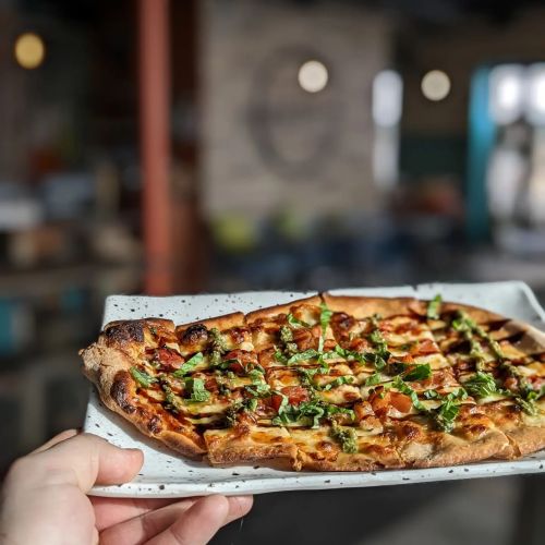 A hand holds a rectangular pizza topped with tomatoes and herbs, set against a blurred indoor background.
