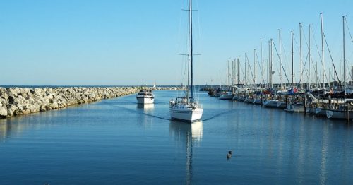Two sailboats navigate a calm marina with a rocky breakwater under a clear blue sky.