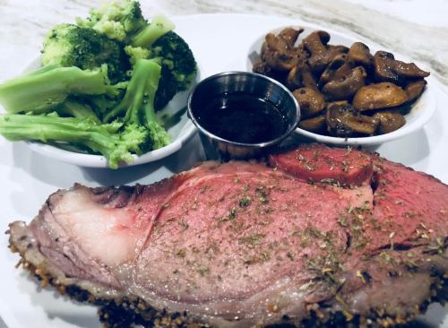 A plate featuring a slice of prime rib, broccoli, mushrooms, and a small cup of sauce.