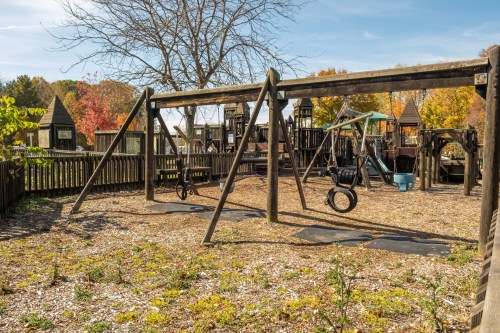 A playground with swings and wooden structures, surrounded by trees with autumn foliage under a clear blue sky.
