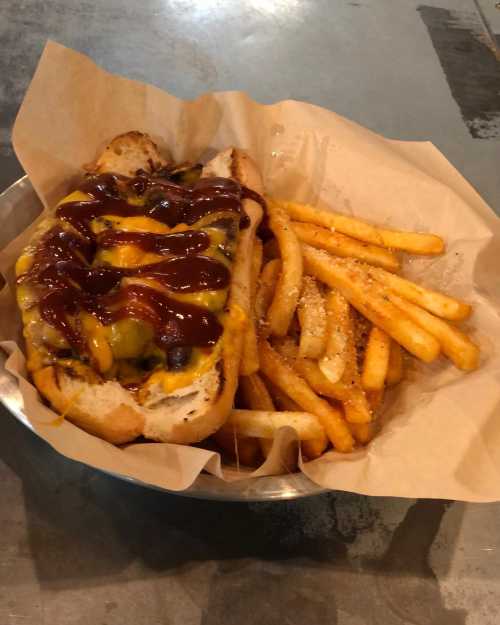 A hot dog topped with cheese and barbecue sauce, served with a side of crispy French fries in a paper-lined bowl.