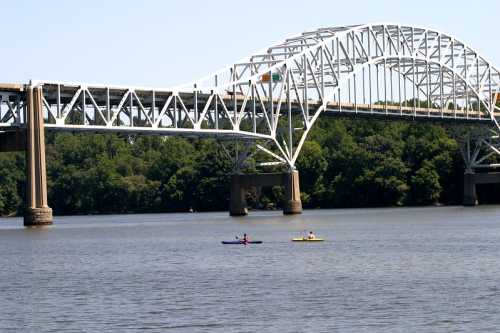 Two kayakers paddle on a river beneath a large white bridge surrounded by green trees.