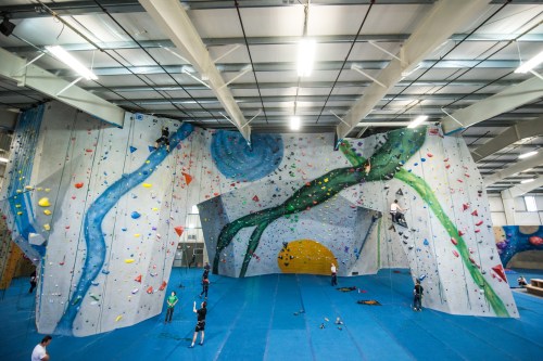 Indoor climbing gym with colorful walls, climbers scaling routes, and a blue mat covering the floor.