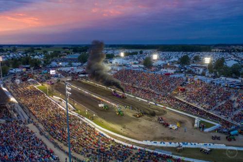 Aerial view of a crowded fairground during a tractor pull event at sunset, with smoke rising and bright stadium lights.