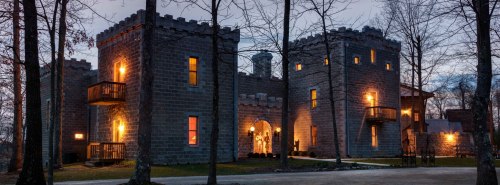 A stone castle with turrets, illuminated windows, surrounded by trees at dusk.