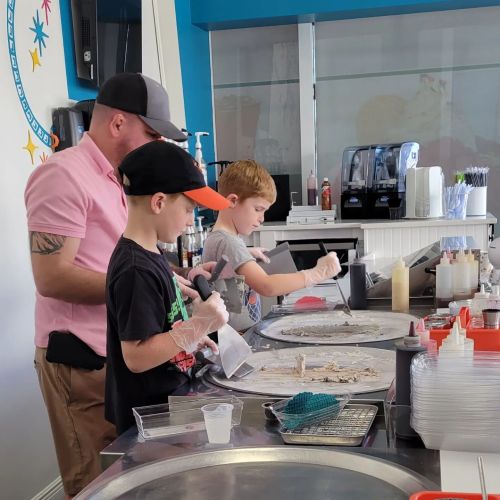 A man and two boys make rolled ice cream at a counter, using spatulas and wearing gloves in a colorful shop.