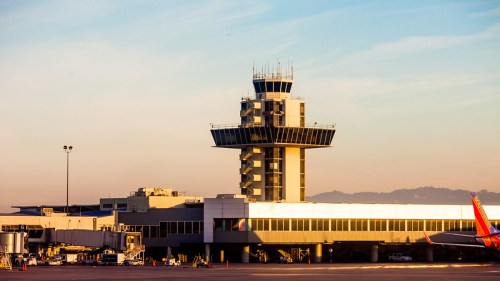A modern airport control tower at sunset, with planes and terminal buildings in the foreground.