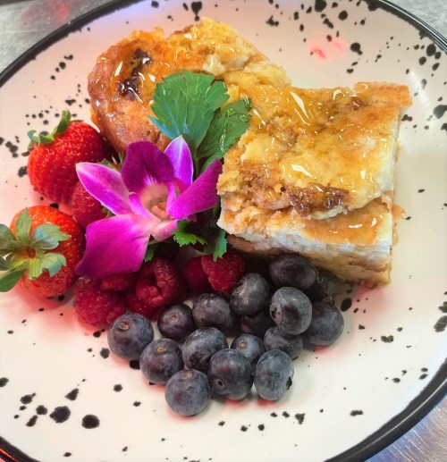A plate with a slice of baked dessert topped with syrup, surrounded by strawberries, raspberries, blueberries, and an orchid.