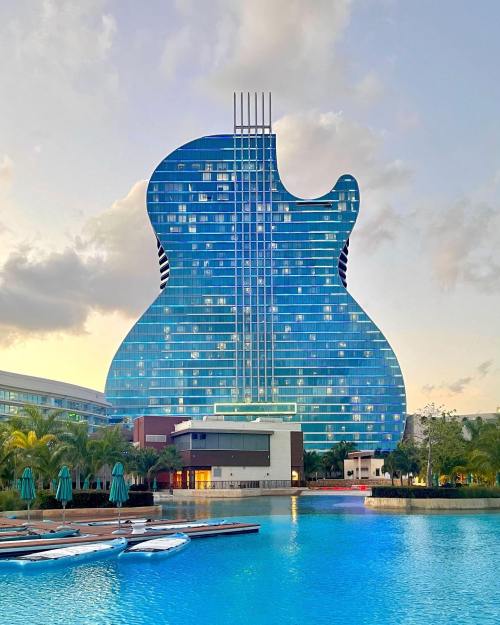 A large guitar-shaped hotel towers over a serene pool, surrounded by palm trees and a clear sky.