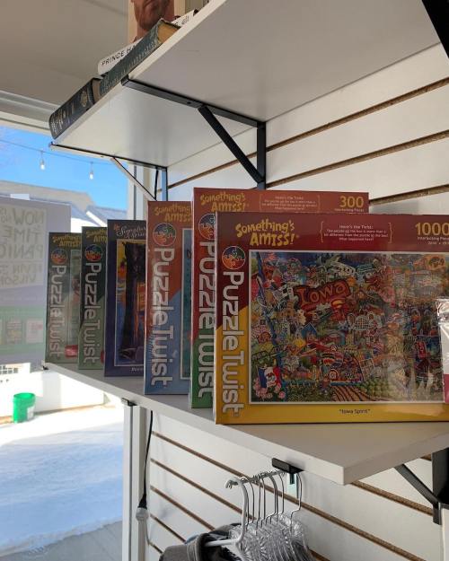 A shelf displaying various jigsaw puzzles, including "Something's Amiss" and "Iowa Spirit," in a bright store setting.
