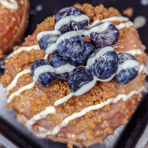 Close-up of a donut topped with blueberries, crumb topping, and drizzled with white icing.