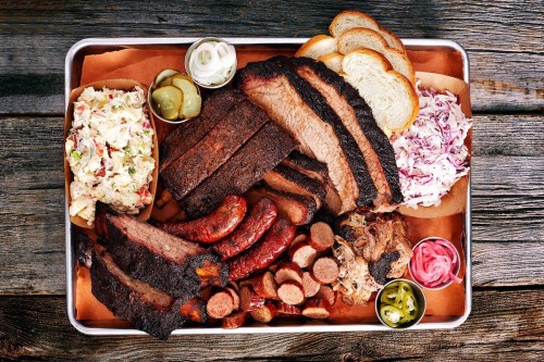 A platter of assorted meats including brisket, sausage, and pulled pork, accompanied by sides of coleslaw, potato salad, and bread.