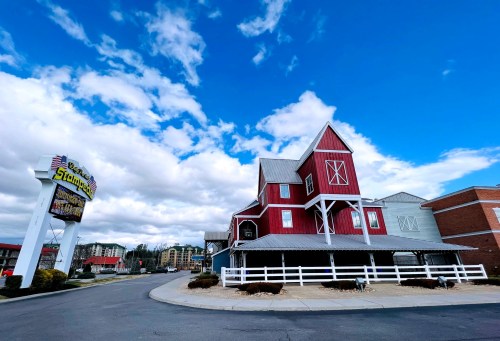 A red barn-style building with a white fence, under a blue sky with clouds, near a roadside sign.