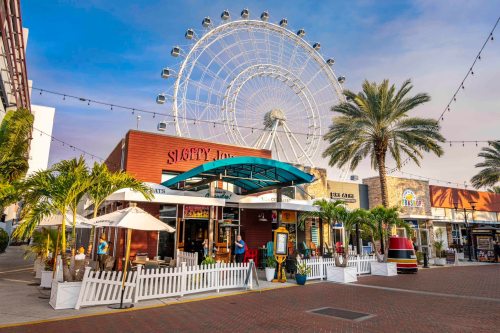 A vibrant outdoor shopping area with palm trees, a restaurant, and a large Ferris wheel in the background.