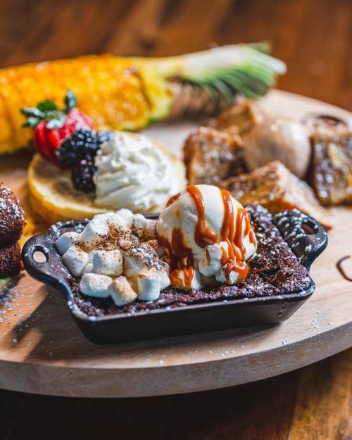 A dessert platter featuring a brownie topped with ice cream and caramel, alongside fruits and other sweet treats.