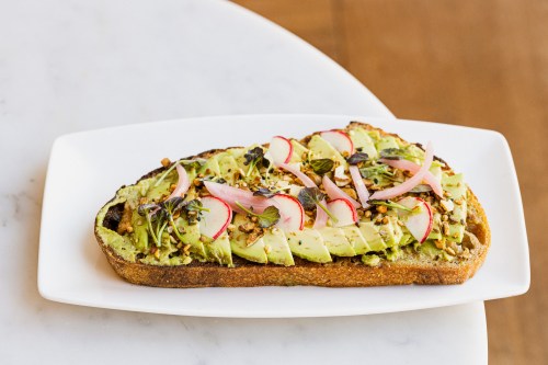 A slice of toasted bread topped with avocado, radishes, pickled onions, and microgreens on a white plate.