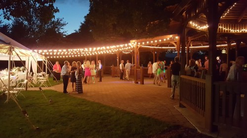 A lively outdoor event with people dancing under string lights, surrounded by tents and a gazebo at dusk.
