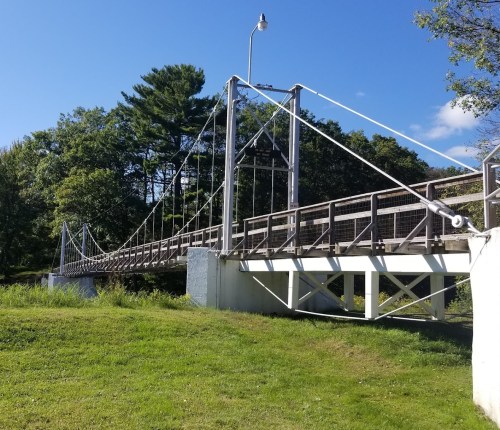 A suspension bridge spans a grassy area, surrounded by trees under a clear blue sky.