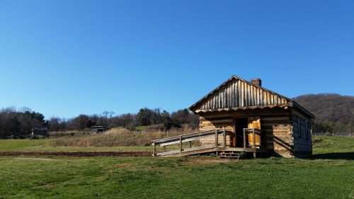 A rustic log cabin with a wooden ramp, set against a clear blue sky and rolling green hills.