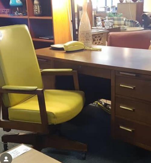 A vintage office setup featuring a yellow chair, a rotary phone, and a wooden desk with drawers.