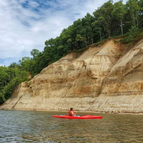 A person in a red kayak paddles along a calm river, with steep, tree-covered cliffs in the background.