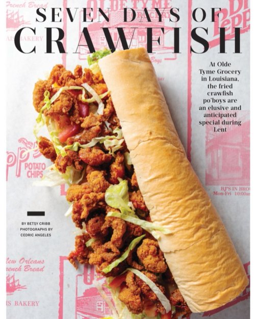 A large crawfish po'boy sandwich filled with fried crawfish, lettuce, and tomatoes on a red patterned background.